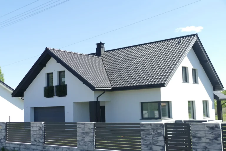 Szary dach dwuspadowy nad białym domem z ogrodzeniem